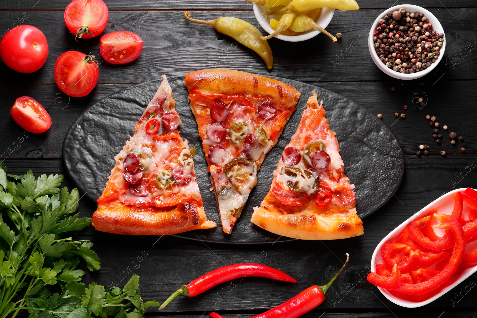 Photo of Slate board with pieces of delicious pizza Diablo among products on black wooden table, flat lay