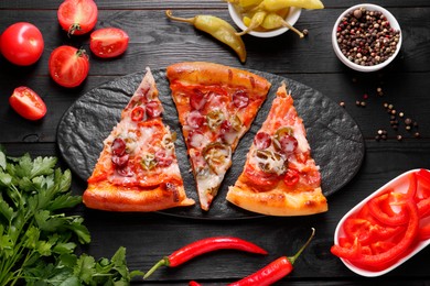 Slate board with pieces of delicious pizza Diablo among products on black wooden table, flat lay