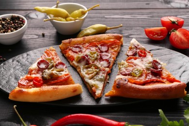 Slate board with pieces of delicious pizza Diablo among products on black wooden table, closeup