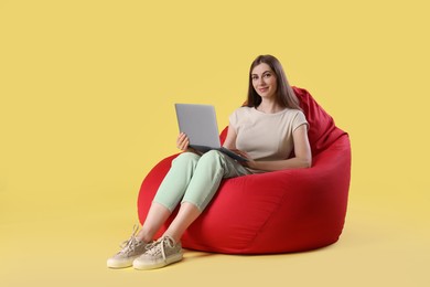 Photo of Beautiful young woman with laptop on red bean bag chair against yellow background