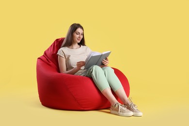 Photo of Beautiful young woman with book sitting on red bean bag chair against yellow background