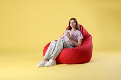 Photo of Beautiful young woman with paper cup of drink sitting on red bean bag chair against yellow background, space for text