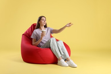 Photo of Smiling woman with paper cup of drink sitting on red bean bag chair against yellow background, space for text