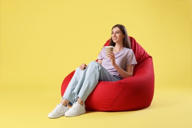 Photo of Smiling woman with paper cup of drink sitting on red bean bag chair against yellow background