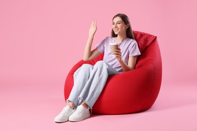 Photo of Smiling woman with paper cup of drink sitting on red bean bag chair against pink background