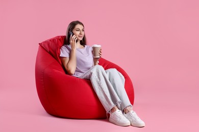 Photo of Beautiful young woman with paper cup of drink talking on smartphone while sitting red bean bag chair against pink background