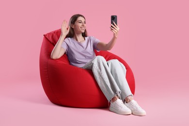 Photo of Smiling woman with smartphone having online meeting on red bean bag chair against pink background