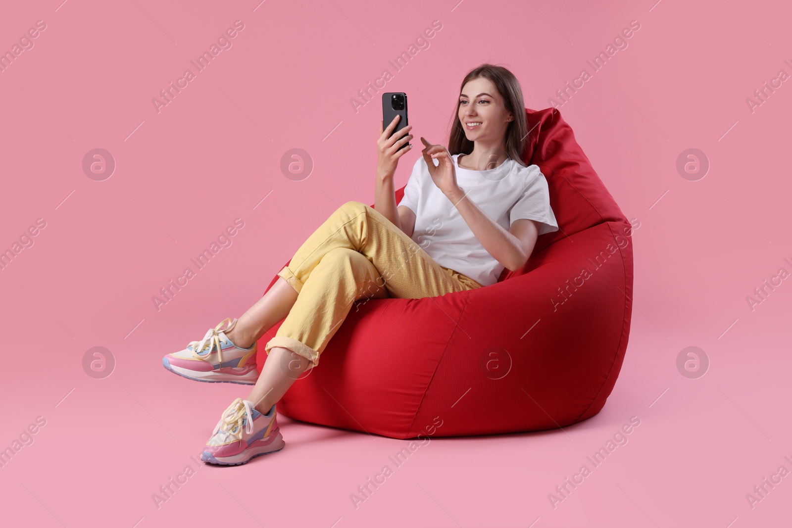 Photo of Smiling woman with smartphone having online meeting on red bean bag chair against pink background