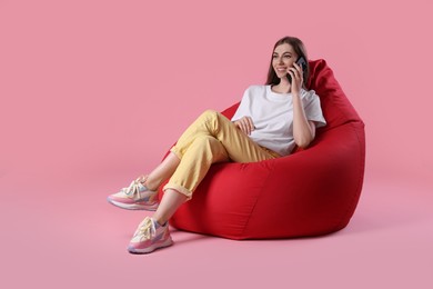 Photo of Smiling woman talking on smartphone while sitting on red bean bag chair against pink background