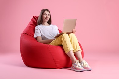 Photo of Smiling woman using laptop on red bean bag chair against pink background