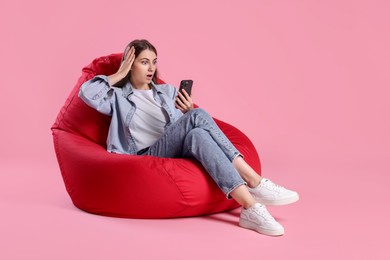 Photo of Emotional woman using smartphone on red bean bag chair against pink background