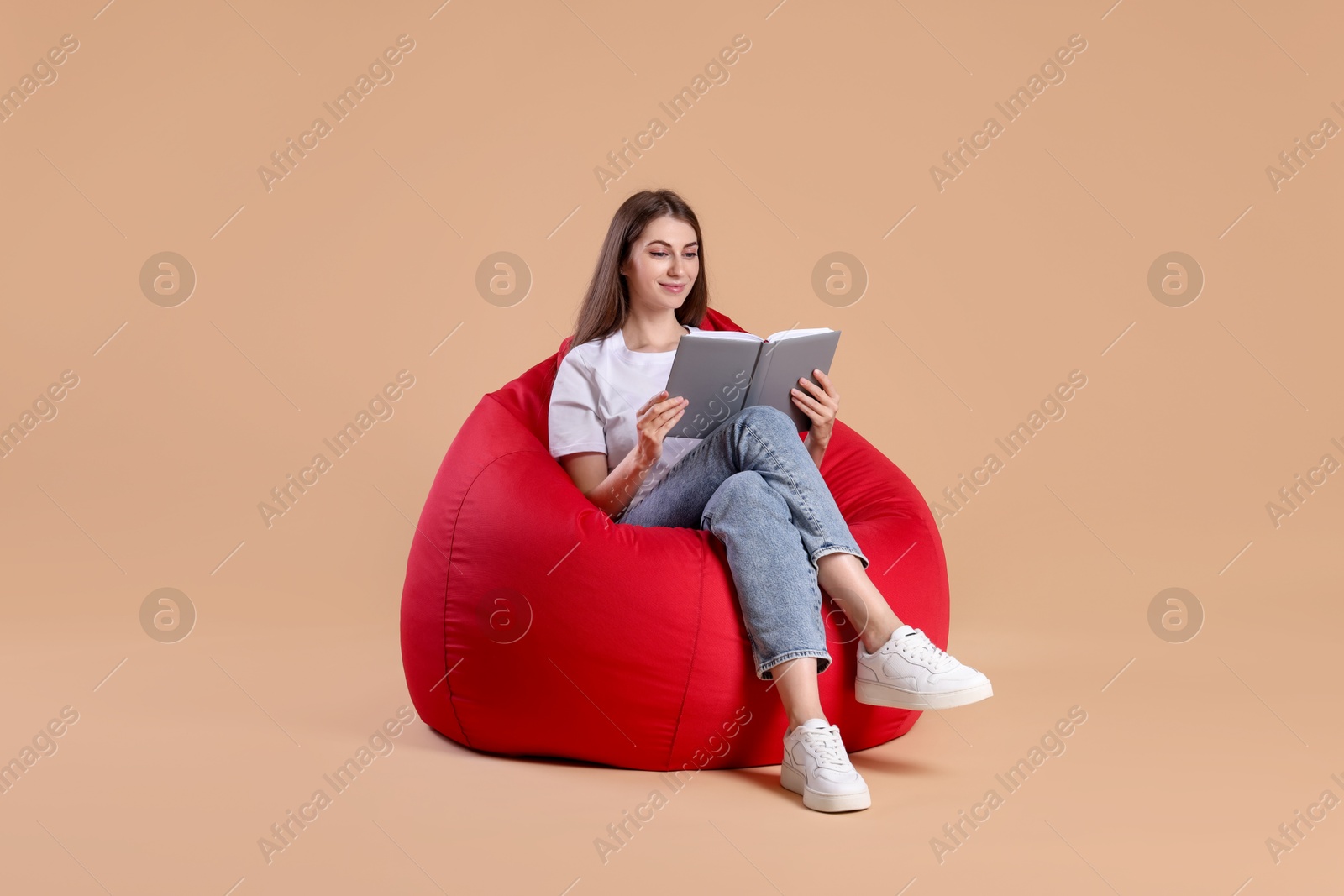 Photo of Beautiful young woman reading book on red bean bag chair against beige background