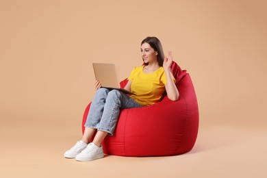 Photo of Smiling woman with laptop having online meeting while sitting on red bean bag chair against beige background