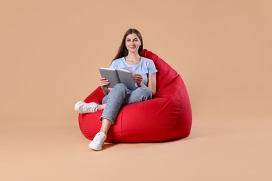 Photo of Beautiful young woman with book on red bean bag chair against beige background
