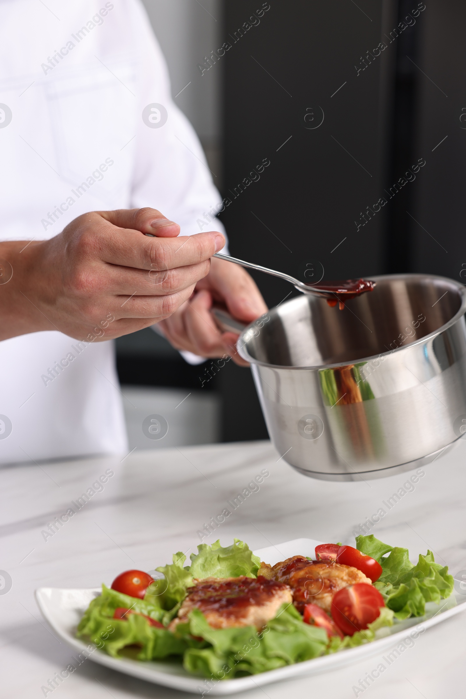 Photo of Professional chef holding saucepan with sauce near delicious dish at white marble table in kitchen, closeup