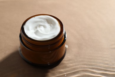 Jar with face cream in water on beige background, closeup. Space for text