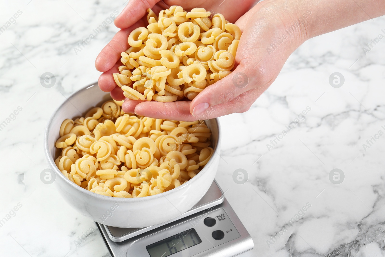 Photo of Woman adding pasta into bowl on kitchen scale on white marble table, closeup