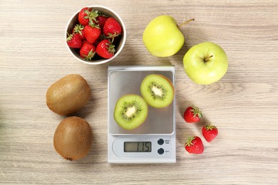 Kitchen scale with halves of kiwi among apples and strawberries on light wooden table, flat lay