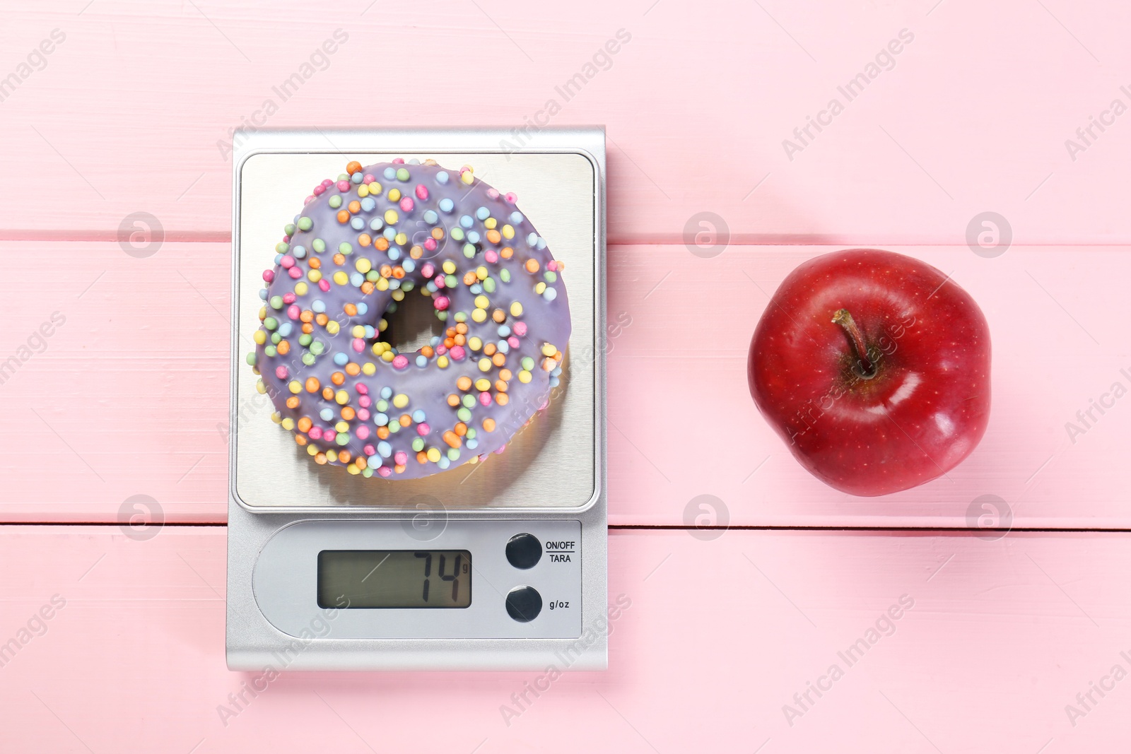 Photo of Kitchen scale with donut and apple on pink wooden table, top view