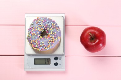 Kitchen scale with donut and apple on pink wooden table, top view