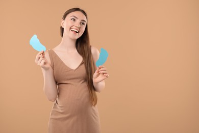 Photo of Expecting twins. Pregnant woman holding two paper cutouts of feet on light brown background, space for text