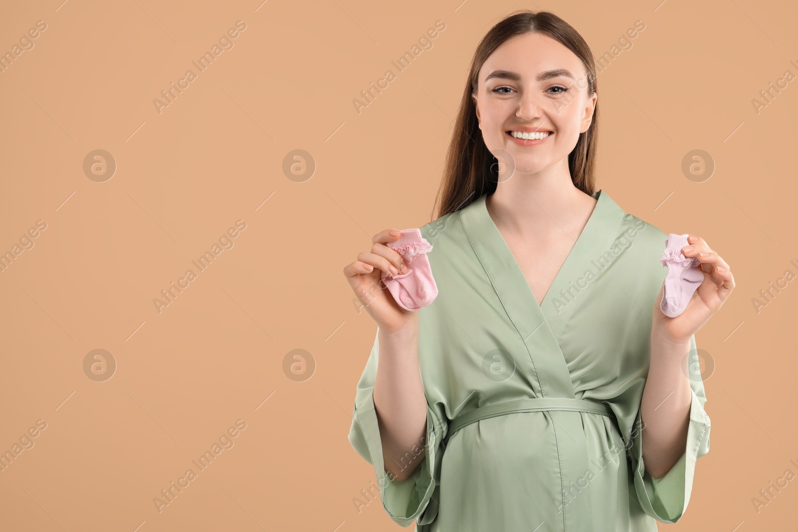 Photo of Expecting twins. Pregnant woman holding two pairs of socks on light brown background, space for text