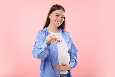 Photo of Expecting twins. Pregnant woman holding two pacifiers on pink background