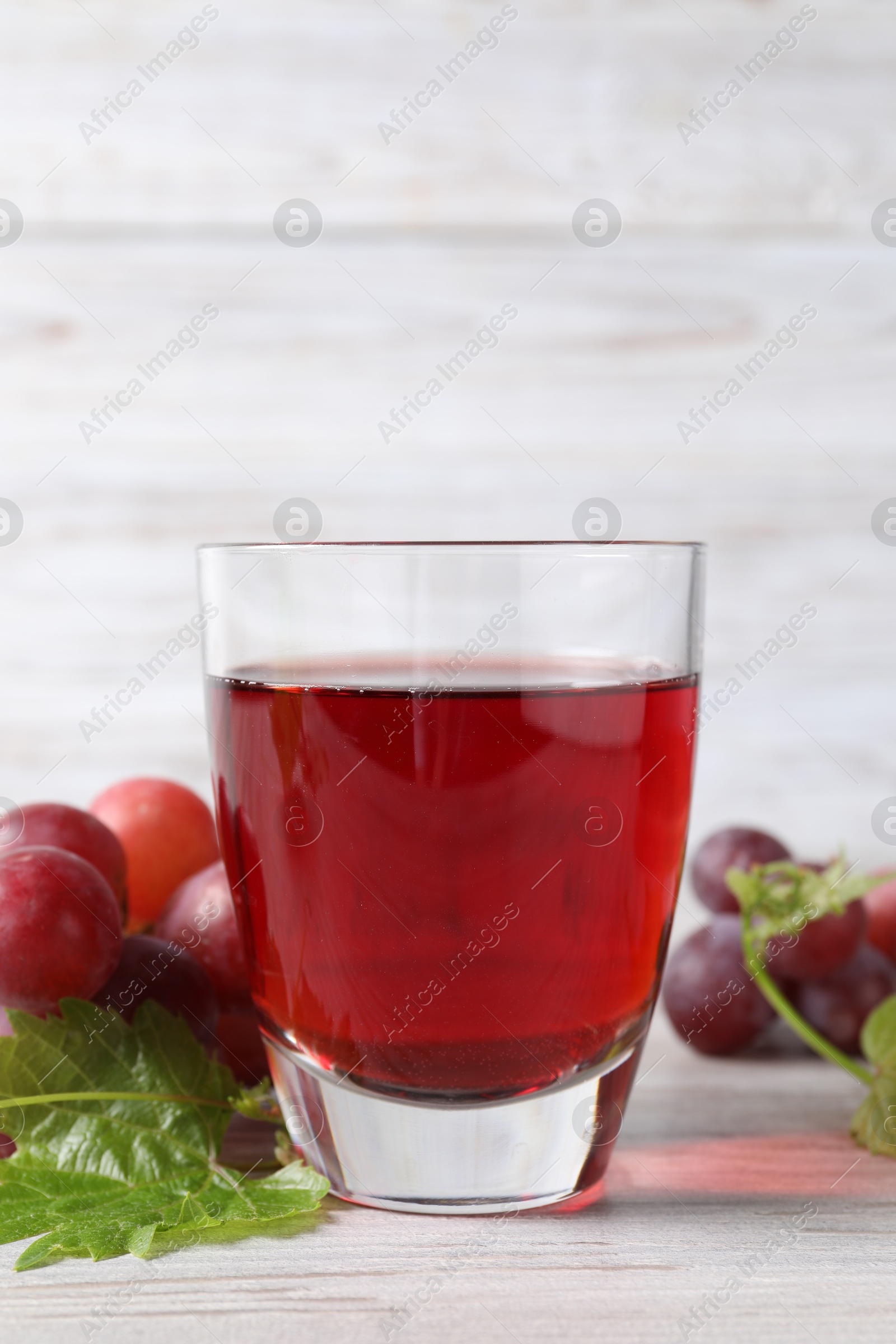 Photo of Tasty grape juice in glass, leaves and berries on light wooden table, closeup