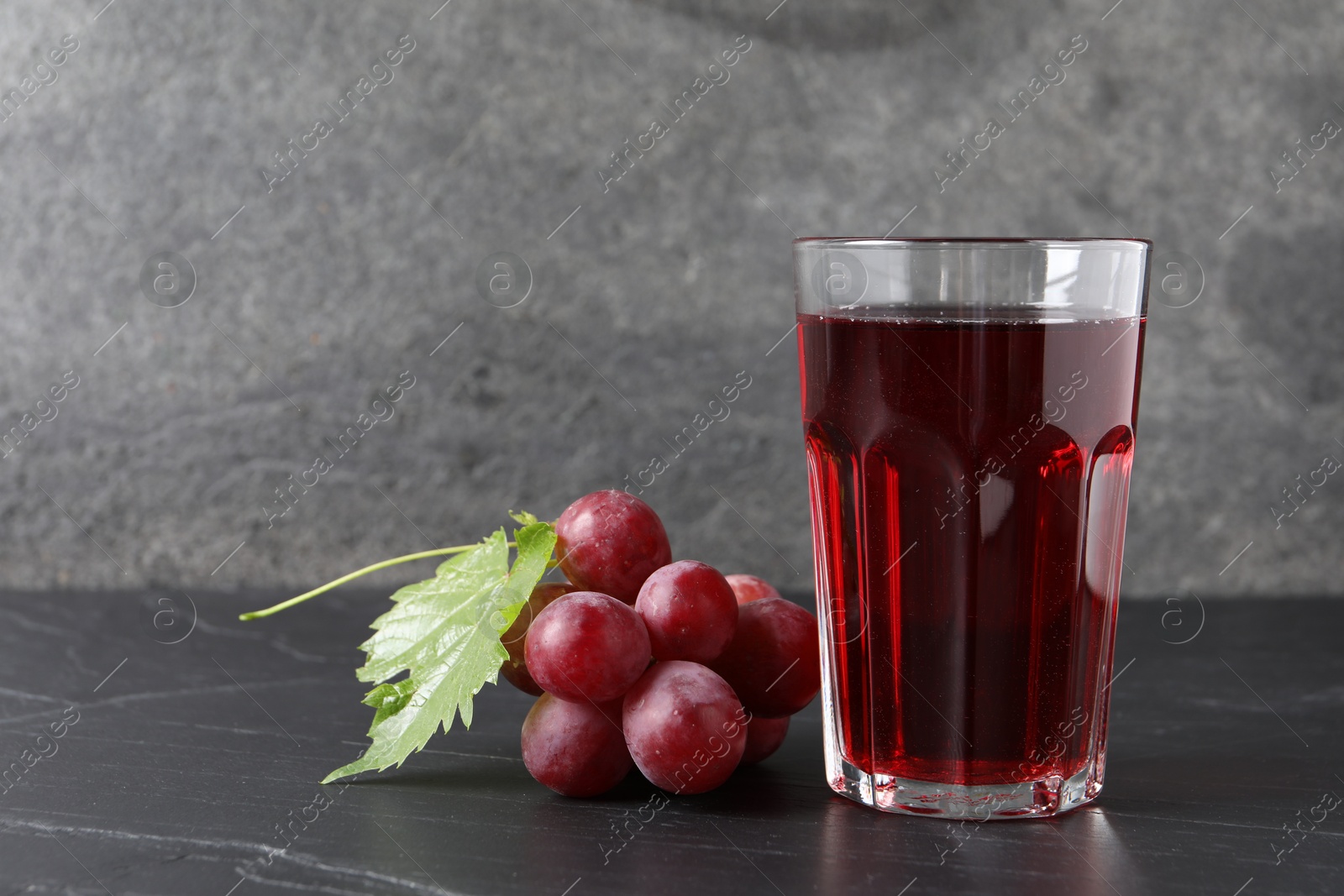 Photo of Tasty grape juice in glass, leaf and berries on dark textured table, closeup. Space for text