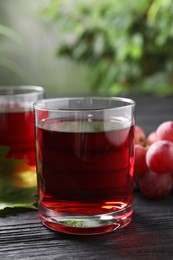 Tasty grape juice in glass and berries on black wooden table against blurred green background, closeup