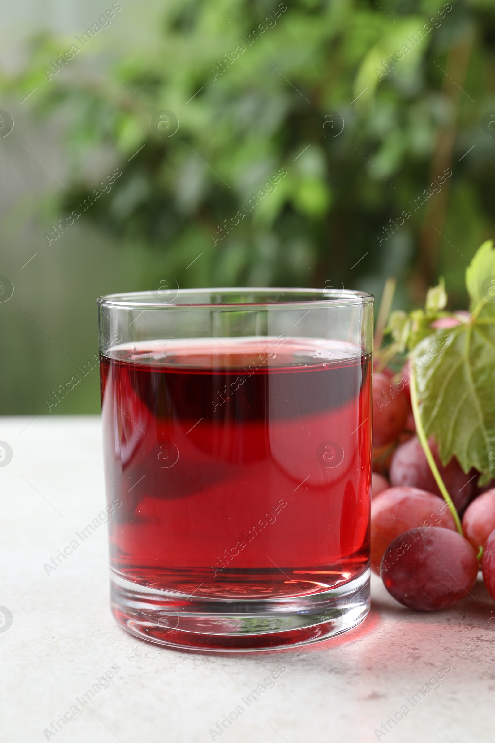 Photo of Tasty grape juice in glass, leaf and berries on light textured table against blurred green background, closeup