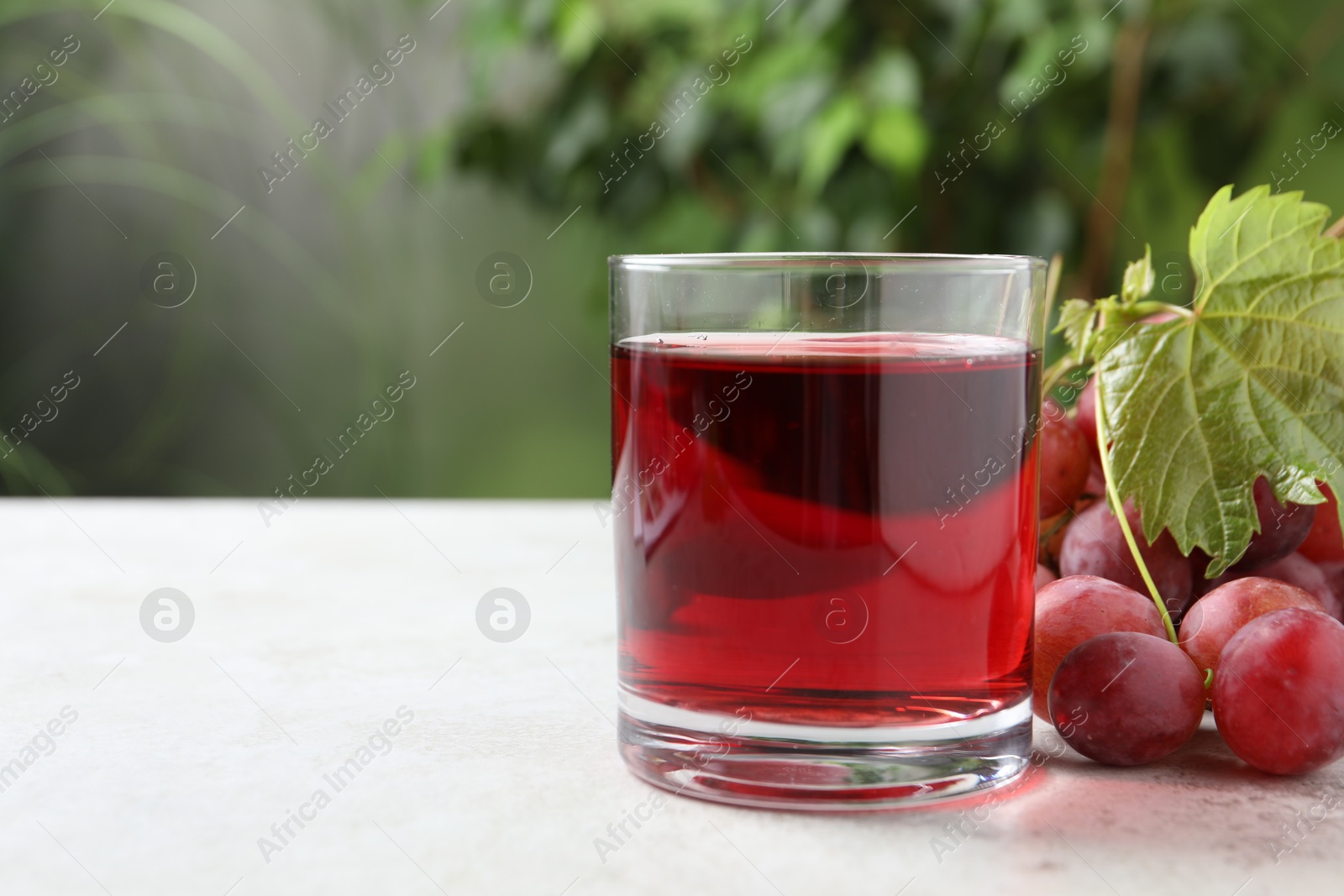 Photo of Tasty grape juice in glass, leaf and berries on light table against blurred green background, closeup. Space for text