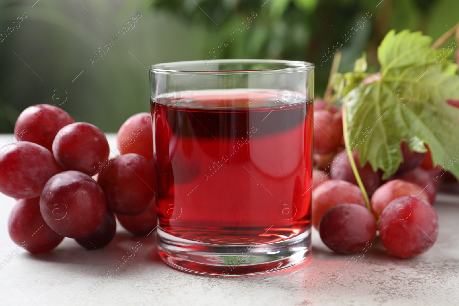 Photo of Tasty grape juice in glass, leaf and berries on light textured table against blurred green background, closeup