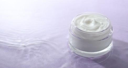 Jar with face cream in water on light violet background, space for text