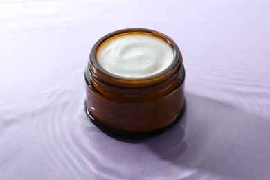 Jar with face cream in water on light violet background