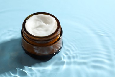 Jar with face cream in water on light blue background, space for text