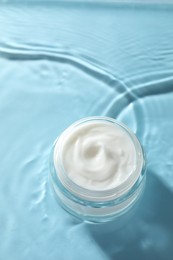 Photo of Jar with face cream in water on light blue background, above view