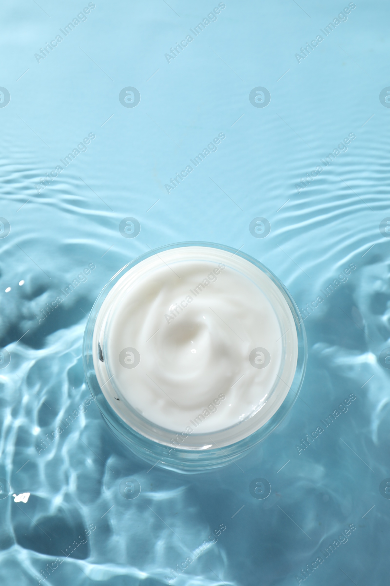 Photo of Jar with face cream in water on light blue background, top view