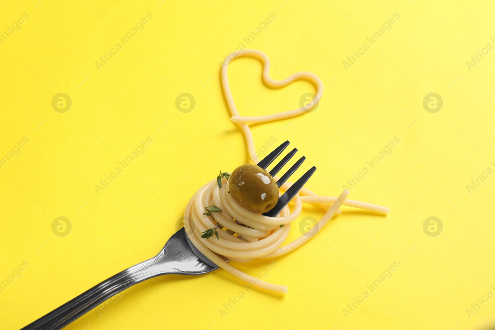 Photo of Heart made of tasty spaghetti, fork and olive on yellow background, closeup