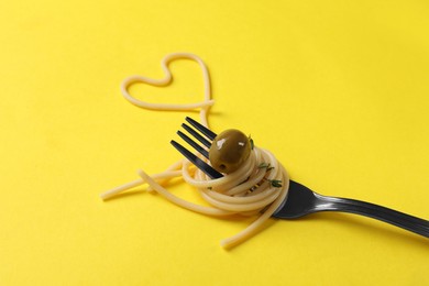 Heart made of tasty spaghetti, fork and olive on yellow background, closeup