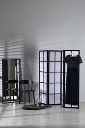 Photo of Folding screen, dress, shoes, chair and mirror near white wall indoors