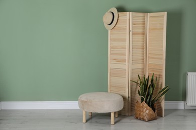 Photo of Wooden folding screen, pouffe, bag, houseplant and hat near green wall indoors, space for text