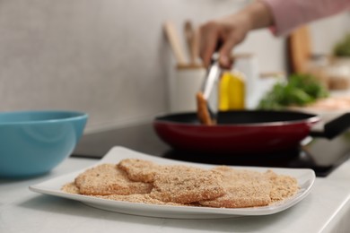 Woman cooking schnitzels in frying pan on stove, focus on raw meat with breadcrumbs