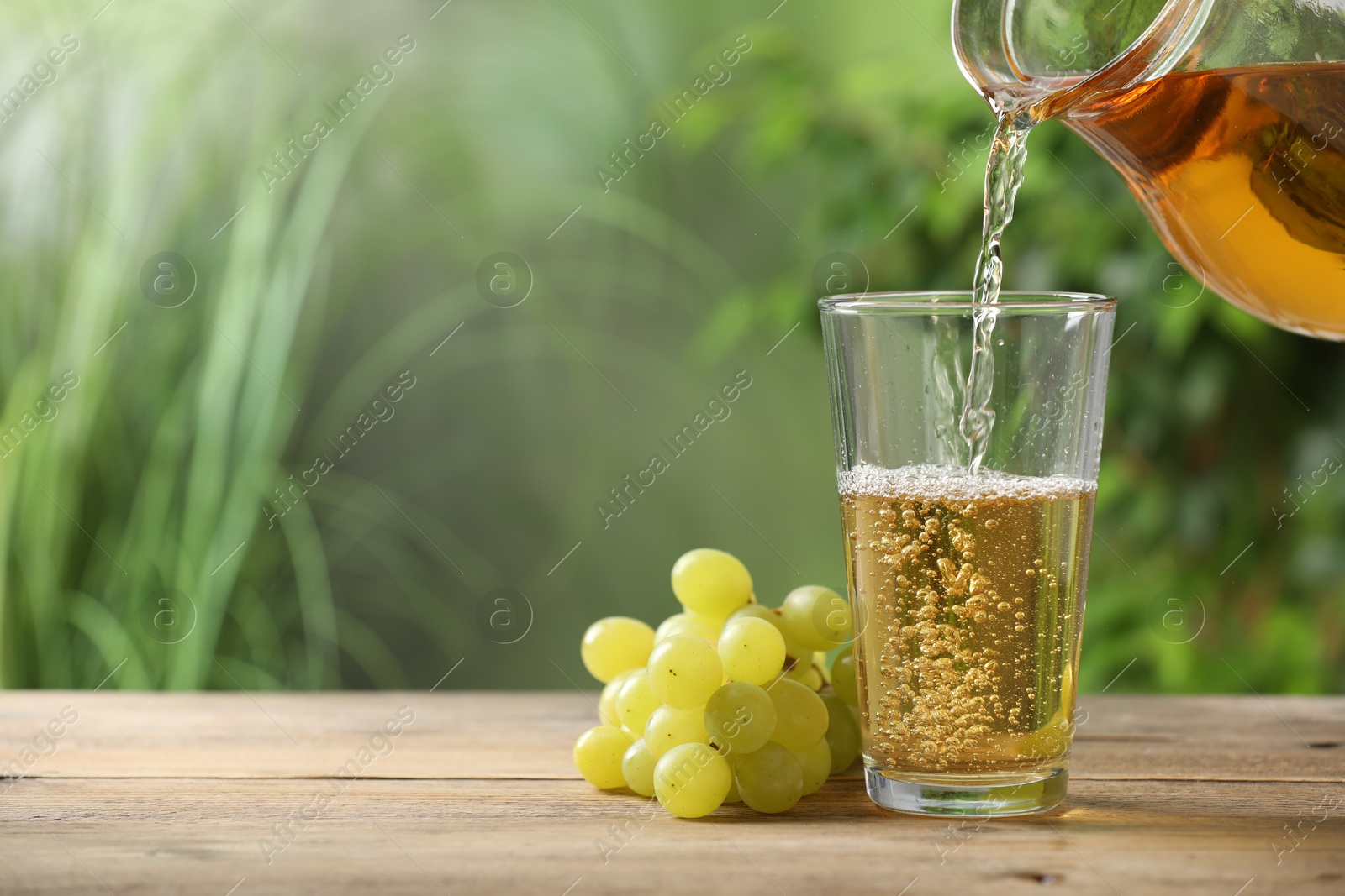 Photo of Pouring tasty grape juice into glass at wooden table outdoors, closeup. Space for text