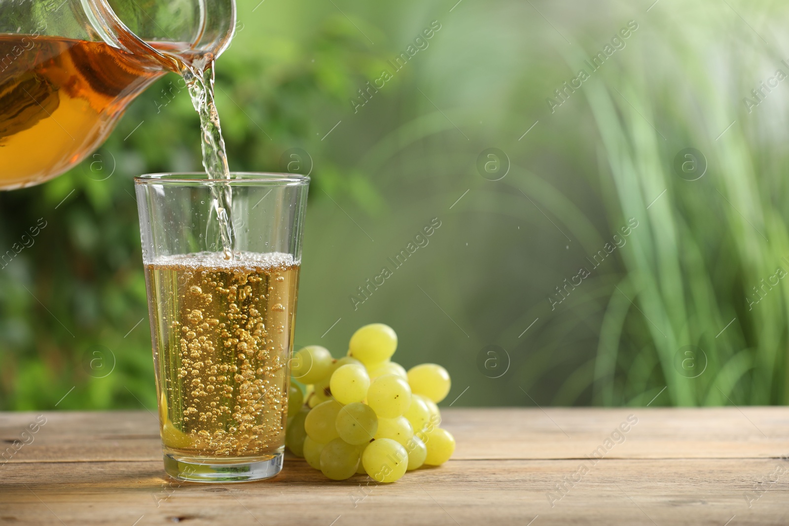 Photo of Pouring tasty grape juice into glass at wooden table outdoors, closeup. Space for text