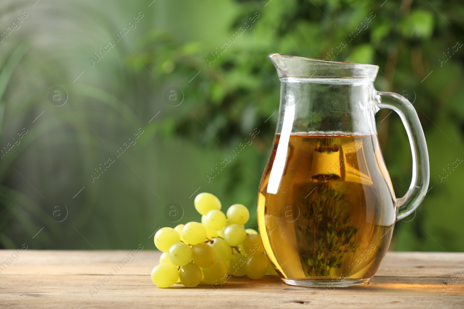 Photo of Tasty juice in glass jug and grapes on wooden table outdoors, space for text