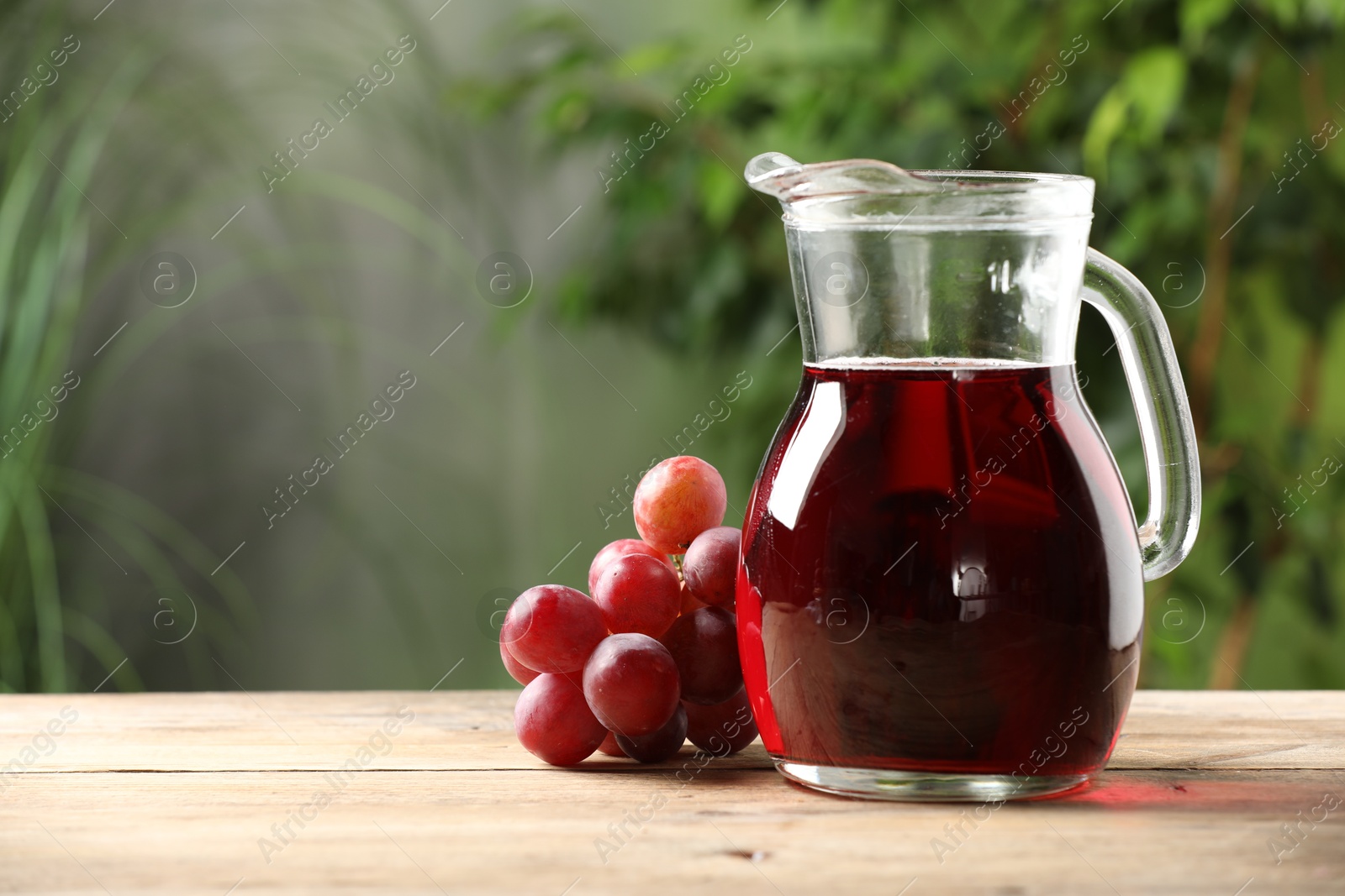 Photo of Tasty juice in glass jug and grapes on wooden table outdoors, space for text