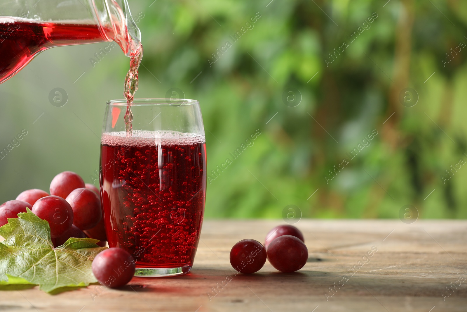 Photo of Pouring tasty grape juice into glass at wooden table outdoors, closeup. Space for text