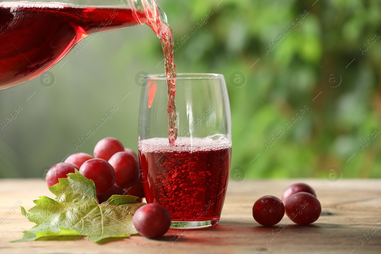 Photo of Pouring tasty grape juice into glass at wooden table outdoors, closeup