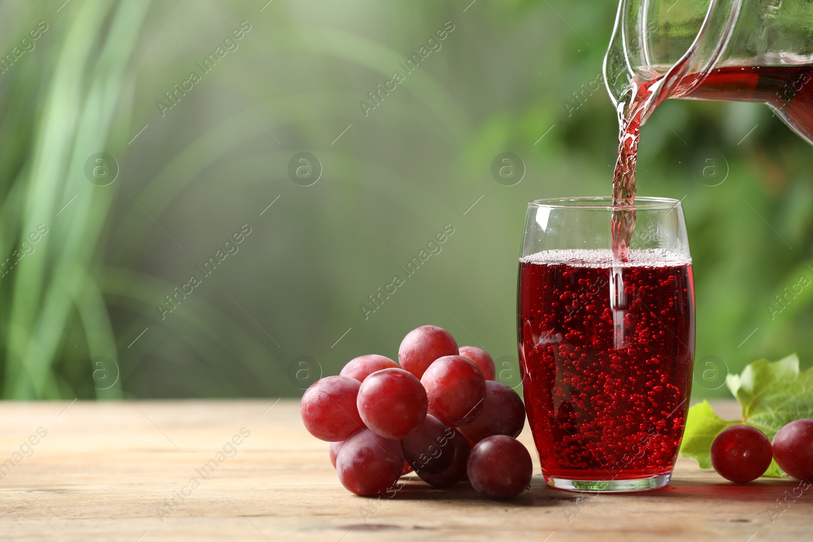 Photo of Pouring tasty grape juice into glass at wooden table outdoors, closeup. Space for text